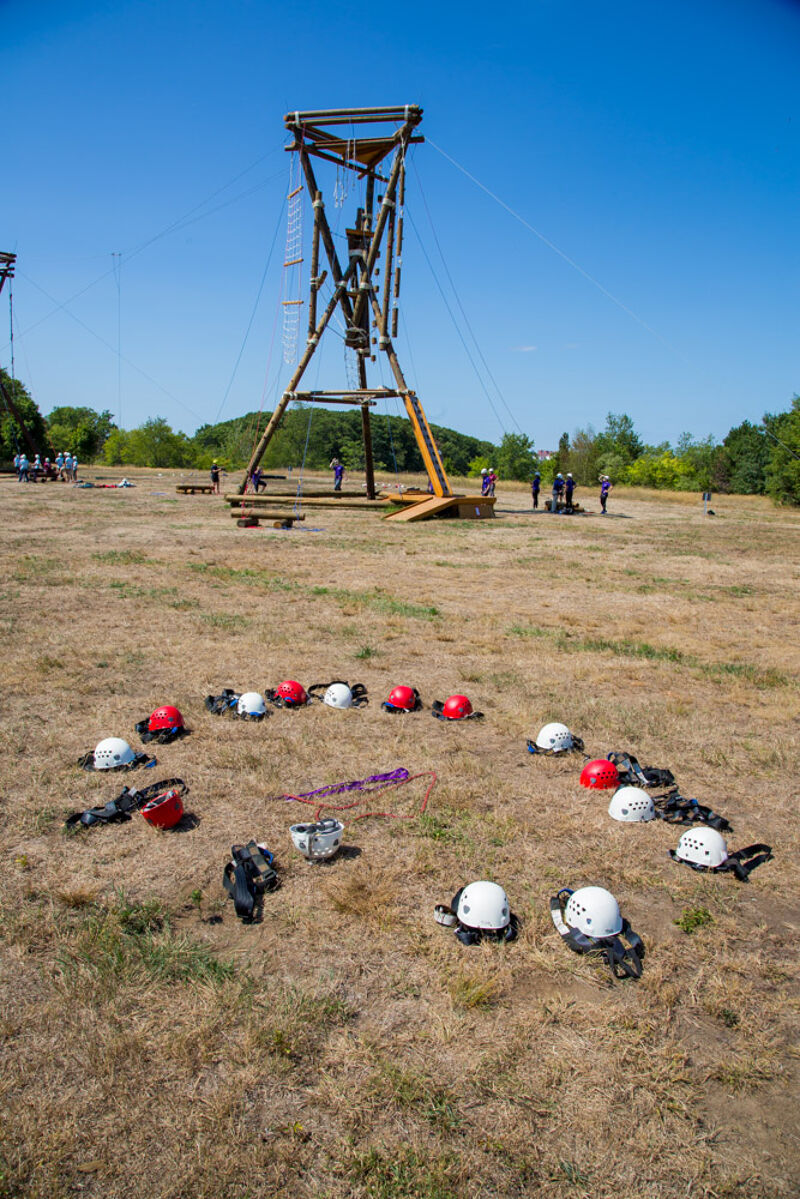 The image shows an outdoor adventure setup on a sunny day. A tall wooden structure with ropes and a ladder stands in the background, likely for activities like zip-lining or climbing. In the foreground, a collection of helmets and harnesses are arranged on the ground, suggesting preparation for an activity. People are visible in the distance.